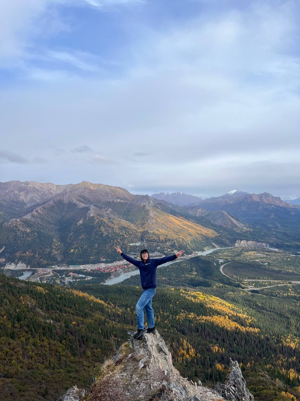Shan Yuxuan standing on a rocky summit with arms outstretched above an autumn valley in Denali, Alaska
